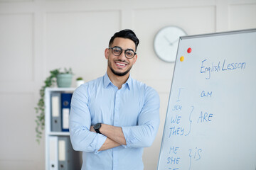 Obraz premium Distance learning. Happy Arab male teacher giving online English class, standing with crossed arms near blackboard, smiling at camera in home office. College tutor explaining grammar rules to students