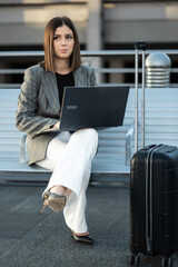 Young businesswoman sitting on a bench, typing on her laptop while her black suitcase waits beside her, embodying remote work and travel convenience in a professional setting