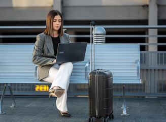 Businesswoman focused on her laptop while sitting on a bench next to a suitcase, representing remote work, business travel, and connectivity during transit