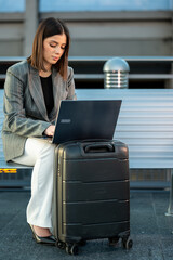 Business woman sitting on a bench, typing on a laptop with her suitcase beside her, embodying the modern concept of remote work and digital nomadism while traveling