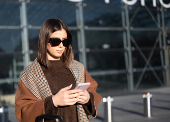 Stylish woman wearing sunglasses and a patterned scarf, standing outside a modern airport building, checking her smartphone while preparing for departure, embodying travel and digital connection