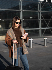 Young woman standing outdoors in front of a modern building, checking her smartphone while pulling a black suitcase, representing urban mobility, connectivity, and modern travel