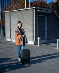 Young woman standing on a city street, pulling a black hard-shell suitcase on wheels, preparing for a journey or arriving at a destination, enjoying travel and leisure in an urban outdoor setting