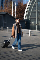 Young adult woman pulling a rolling suitcase, walking on a paved pathway near modern architecture and bare trees, representing travel, departure, and urban mobility concepts
