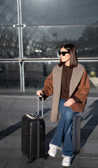 Stylish young woman wearing sunglasses, a brown sweater, and a brown checkered jacket, sitting with her black rolling luggage, ready for departure from a station or airport terminal building