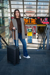 Young woman standing on a platform holding a black suitcase, ready for a journey at a modern train station with a departure board in the background, representing travel and transportation concepts