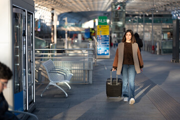 Female traveler pulling wheeled suitcase through a bright, modern train station terminal, walking toward departure or arrival amid blurred commuters, urban travel lifestyle scene