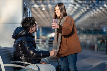Woman standing and confronting man sitting on a bench, having an intense discussion with angry expressions, pointing fingers, and holding a phone at a modern transportation hub