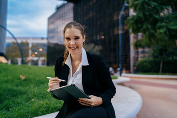 Smiling businesswoman using tablet and stylus in city park, expressing enjoyment of mobile creative task, balancing professionalism and artistic energy in digital workspace