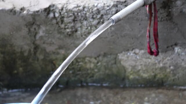 Clean mountain water flows steadily from a pipe near a wall, showing a simple rural water supply system used for drinking and daily needs in Uttarakhand, India.