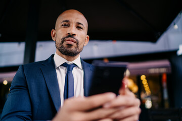 Businessman holding smartphone with serious look, making direct eye contact, symbolizing confidence, digital authority and assertive presence in professional tech-based environment.