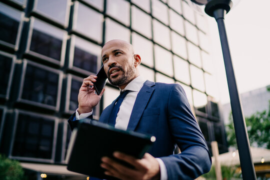 Professional businessman communicating on smartphone and holding tablet, speaking assertively, embodying mobile control, leadership and fast decision-making in corporate tech world.