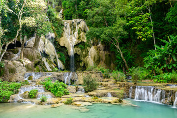 Vivid green forest surrounds tiered limestone cascades and clear turquoise pools at Kuang Si Falls in northern Laos. The scene features lush foliage, layered rock formations, and flowing water in a © Florent