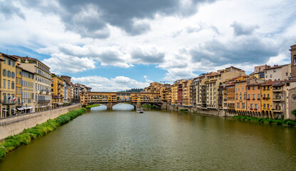 Fototapeta premium View of Ponte Vecchio in Florence (Ponte Vecchio, Firenze) and nearby buildings along the Arno River on a bright summer day, capturing the historic charm and vibrant riverside scene.