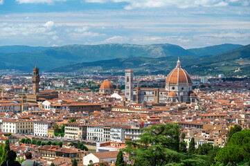 Panoramic view of Florence (Firenze), Italy, showing rooftops, landmarks like Palazzo Vecchio and Santa Maria del Fiore with its red dome, and hills with forests on a sunny summer day.