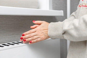 A girl in a knitted sweater warms her hands near a radiator. A girl warms herself near a heating radiator. heating radiator. person warming up. 