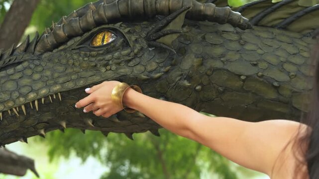 female hand stroking fantasy dragon, muzzle head close-up, ancient monster dinosaur, dark green reptile. Background forest trees nature summer. Girl queen of dragons fairy world. Gothic princess woman