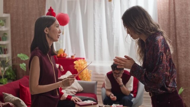Side view of two young Asian girlfriends sharing red envelopes with greetings during traditional indoor Tet celebration