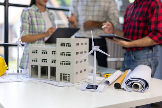 Miniature wind turbine and solar panel models are placed on a table alongside blueprints and a building model, while architects discuss a sustainable building project in the background. - Powered by Adobe
