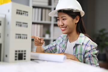 Young Asian female architect wearing a safety helmet is sitting at her desk, pointing at a building model and smiling.