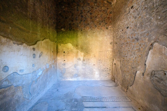 Ancient Roman room in the House of Menander, Pompeii, Italy, with textured stone walls, faded plaster, and a geometric mosaic border on the floor. Soft daylight highlights the rustic surfaces and