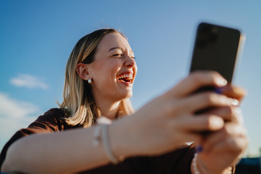 Happy young woman laughing while taking selfie with smartphone outdoors, enjoying digital communication, mobile connection and modern lifestyle in urban environment.