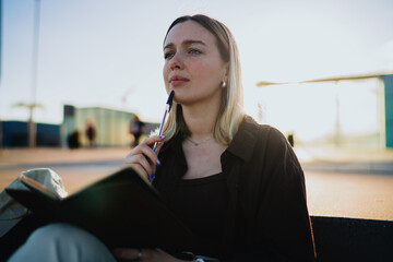 Fototapeta na wymiar Young woman looking thoughtful with notebook and pen outdoors, gazing away with focus, symbolizing reflective thinking, study, education and creative freelance inspiration.