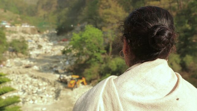 An adult woman stands on a mountain roadside watching traffic on a winding hill road, reflecting travel, safety awareness, and everyday life in Uttarakhand, India.