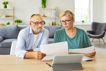 Senior couple sits at table with tablet and paperwork, discussing budget, expenses and retirement...