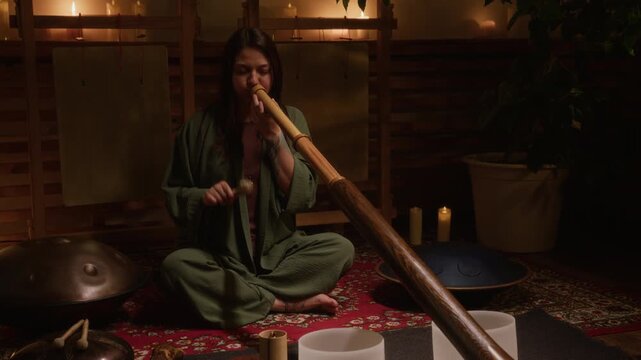 Medium full shot of young Chinese woman lying on carpet with closed eyes, meditating and relaxing during sound healing session, while Caucasian female practitioner is playing didgeridoo with rattle