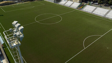 High-angle aerial view of a modern soccer field with dark green artificial turf, crisp white markings, stadium seating, and a large floodlight tower. Aerial shot of a small football stadium.