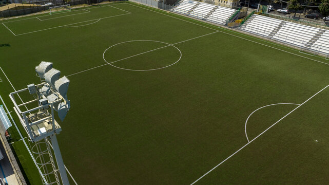 High-angle aerial view of a modern soccer field with dark green artificial turf, crisp white markings, stadium seating, and a large floodlight tower. Aerial shot of a small football stadium.