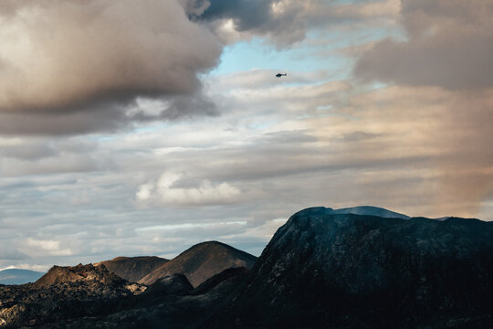 View of dark, rugged mountains rise against a sky swirling with clouds and a hint of blue, while a helicopter flies above, Fagradalsfjall, Reykjanes Peninsula, Iceland.