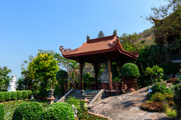 Fototapeta premium Decorative wooden pavilion with intricate carvings stands on a stone platform surrounded by manicured shrubs, lush greenery, and a pond filled with water lilies under a clear blue sky. The peaceful