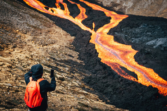 View of a person with a bright orange backpack taking a photo of flowing rivers of fiery orange lava over black rock, Fagradalsfjall, Reykjanes Peninsula, Iceland.