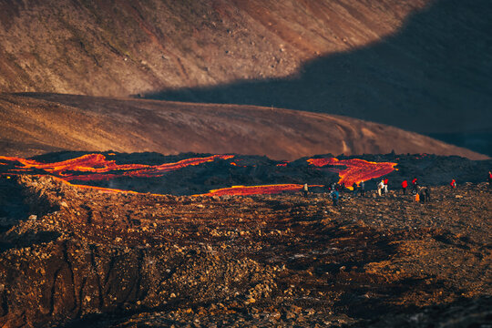 View of fiery orange lava flows across the rugged, dark volcanic landscape as people gather near the Geldingadalir eruption, Fagradalsfjall, Reykjanes Peninsula, Iceland.