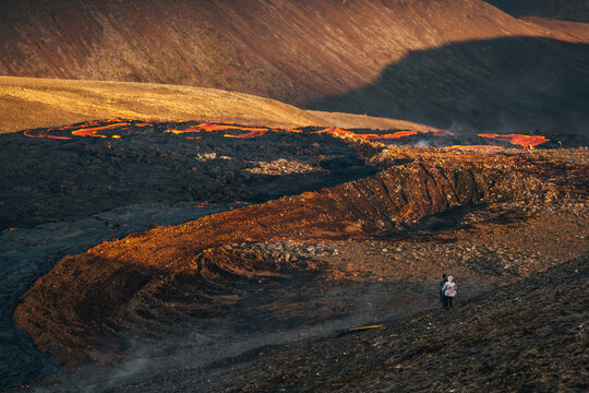 View of a lone figure stands dwarfed by the vast, undulating landscape of cooled lava and fiery rivers, contrasting starkly against the rugged mountains, Fagradalsfjall, Reykjanes Peninsula, Iceland.
