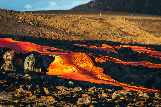 View of a brave photographer with a backpack capturing the fiery river of molten lava snaking through jagged black volcanic rocks, Fagradalsfjall, Reykjanes Peninsula, Iceland.