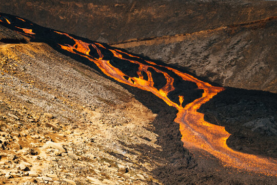 View of molten lava flowing like a fiery river through a desolate landscape of dark rock, contrasting starkly against the rugged terrain, Fagradalsfjall, Reykjanes Peninsula, Iceland.