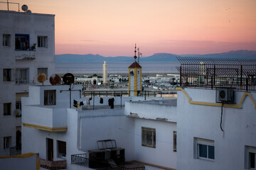 Evening cityscape of Tangier with rooftops, mosque minaret and harbor in the distance, overlooking the sea at sunset under a soft pink sky.