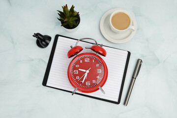 Top view flat lay of red alarm clock on open notebook with pen, coffee cup, plant, and desk lamp....