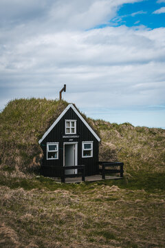 View of a quaint black house with a grass roof nestles into the earth under a cloudy sky, a serene contrast of colors and textures, Saudarkrokur, Drangey Island, Iceland.