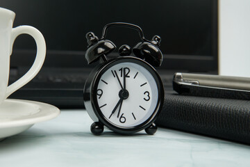 Office workspace with black alarm clock beside laptop, notebook, pen, and coffee cup. Concept of...