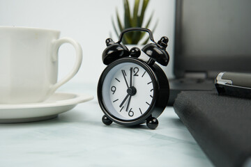Office workspace with black alarm clock beside laptop, notebook, pen, and coffee cup. Concept of...