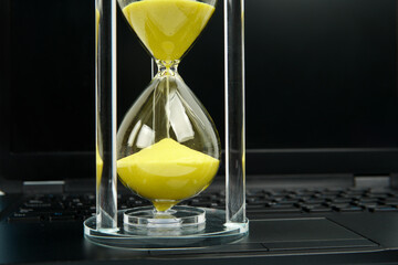 Close up hourglass with yellow sand on laptop keyboard. Concept of time passing, deadline countdown, productivity pressure, efficiency, and professional time management.