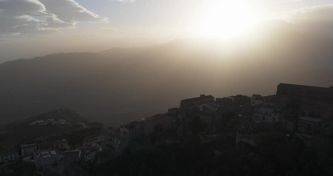 Aerial view of Pollina's landscape, where the silhouette of buildings meets the horizon under a sunlit sky, creating a blend of shadow and light, Pollina, Sicilia, Italy.