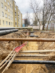 Construction site showing deep trench with various pipes and cables, surrounded by buildings and trees, illustrating urban infrastructure development and maintenance