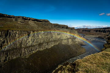 View of a vibrant rainbow arcs over the rugged cliffs and the rushing river below, reflecting in the water near Dettifoss waterfall, Vatnajokull Park, Northeast Iceland, Iceland.