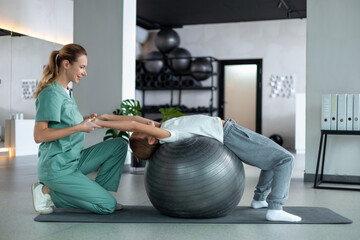 Female therapist and child doing back exercise lying on fitness ball