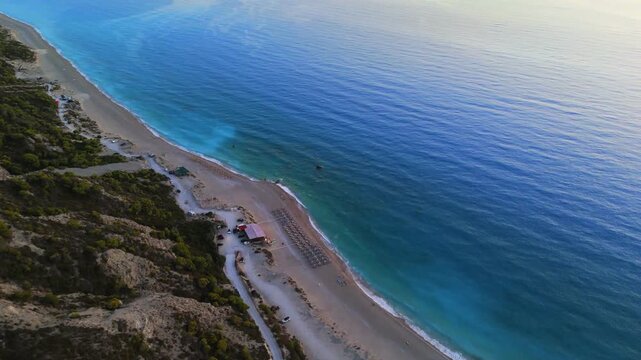 Aerial view of the coastline meeting the turquoise sea, with a beachside structure and umbrellas contrasting against the natural landscape, Lefkada Island, Lefkada, Greece.